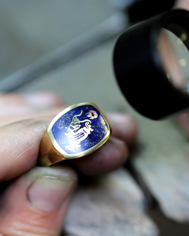 A yellow gold signet ring with blue enamel and a dragon design is being looked at by a jeweller in the workshop through a magnifying glass